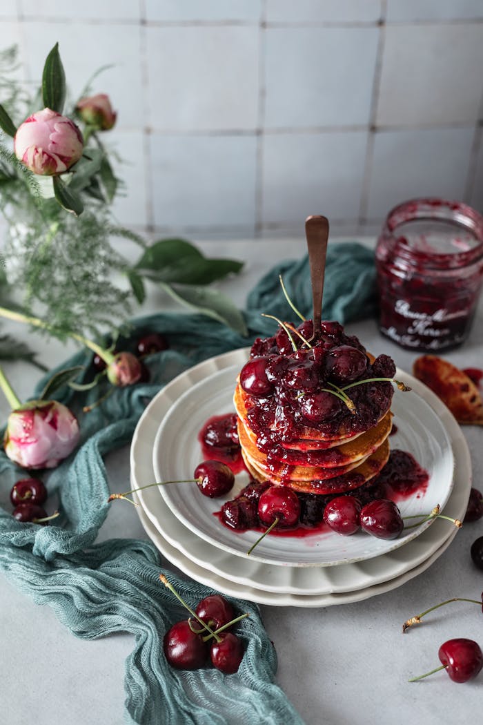 service-02 Homemade pancakes topped with syrupy cherries, surrounded by flowers on a rustic table.